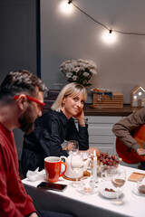 A group of friends sitting at a table with glasses, candles, snacks, and playing guitar, enjoying an intimate and cozy evening together in a relaxed atmosphere.