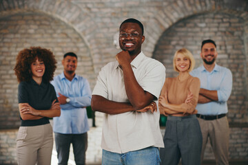 Confident African American businessman stands out in front of his successful and diverse multi-ethnic startup team with arms crossed in a modern office