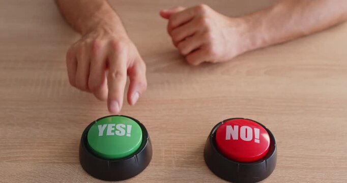 A hand hovers between a green YES button and a red NO button on a desk, illustrating hesitation, decision, choice. Man making decision sitting at table. Future solution, selection and alternative.