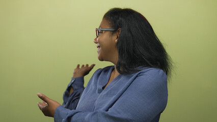 African american woman wearing glasses smiling points hand to side in green studio; invitation warmth.