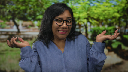 Woman shrugs shoulders in forest clearing wearing blue blouse amid green trees; uncertainty...
