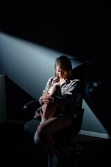 Obraz premium A woman sits contemplatively on a chair, illuminated by a dramatic single light source, creating high contrasts and evoking introspective and meaningful emotions in a minimalist dark environment.