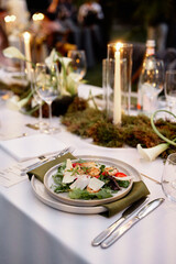 Sophisticated outdoor dinner setup featuring fresh salad with greens and shaved parmesan, lit candles, wine glasses, and greenery centerpiece, warm, festive ambiance for party or wedding reception.
