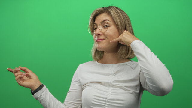 Woman miming phone gesture with hand and pointing finger in studio against green screen, wearing glasses and white shirt; playful. - Powered by Adobe