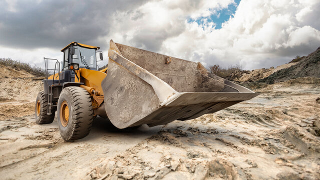 A large excavator loader digs into the sandy terrain at a construction site, surrounded by clouds softly drifting across the blue sky.