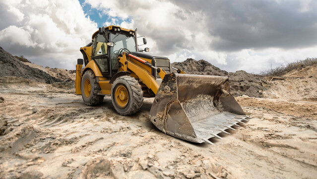 A yellow backhoe loader operates on a gravel surface, moving dirt and debris under an overcast sky, showcasing the power of construction machinery in action