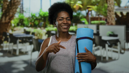 Woman holding yoga mat on a city terrace, surrounded by outdoor restaurant seating, showcasing a blend of fitness and urban life during a warm day.