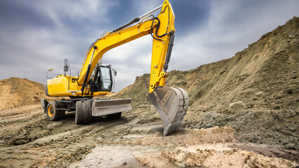 A large yellow excavator digs into the sandy ground at a construction site. Dark clouds loom overhead, creating a dramatic backdrop for the busy work