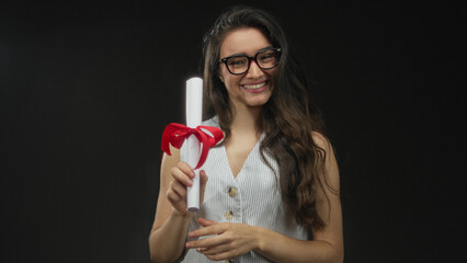 Young woman holding diploma scroll with red ribbon against a black studio backdrop; pride achievement.