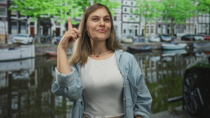 Woman smiling and holding up two index fingers pointing toward canal water on street beside...