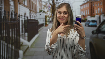 Fototapeta premium Young blonde woman pointing finger to blue bottle while giving thumbs up on urban street; health optimism.
