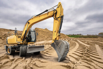 Obraz premium A yellow excavator maneuvers through a construction site, skillfully reshaping the sandy terrain as overcast skies loom above on a quiet afternoon