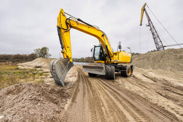Obraz premium Yellow excavator digs into the sandy terrain beside a muddy riverbank, surrounded by earthy mounds, while another crane looms in the distance