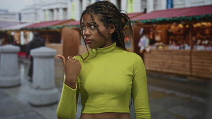 Woman touching braid with hand at street market stall wearing green turtleneck sweater and looking to side; uncertainty reflection.