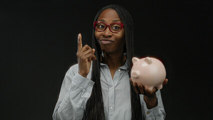 Woman holding pink piggybank wearing glasses against black background, suggesting savings and financial concepts, isolated in studio setting emphasizing calm confidence.