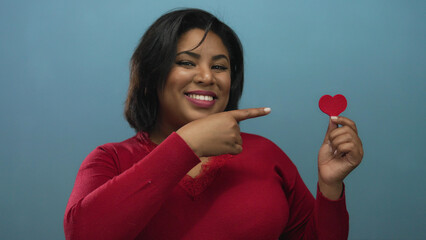 Latina woman in red sweater holding and pointing at a paper heart against a blue background,...