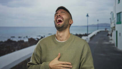 Fototapeta premium Young hispanic man laughing joyfully on seaside promenade with ocean backdrop, showcasing a relaxed outdoor scene filled with happiness and tranquility at the beach.