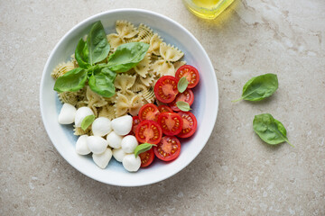 Plate of ingredients for caprese pasta salad with farfalle, horizontal shot on a beige stone background, above view