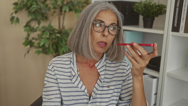 Woman holding red smartphone near mouth and touching neck while speaking in office building with bookshelf and plant visible; worry health call.