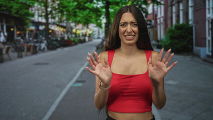 Young woman with hands raised palms out on a city street, red crop top showing midriff and a worried grimace; refusal discomfort.