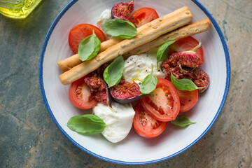 Salad with fresh tomatoes, torn figs, mozzarella, basil and grissini served in a white and blue plate, middle close-up, horizontal shot