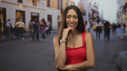 Woman with hand on chin and bare shoulder on crowded city street, smiling in red top, casual pose and watch visible; thoughtful.