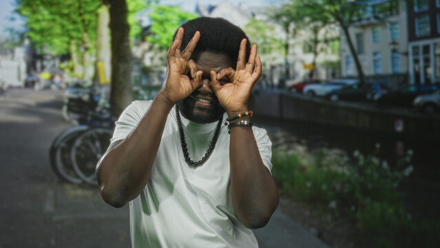 Man making hand binoculars and ok sign on a sunny street by a canal with bicycles and parked cars visible; playful curiosity.