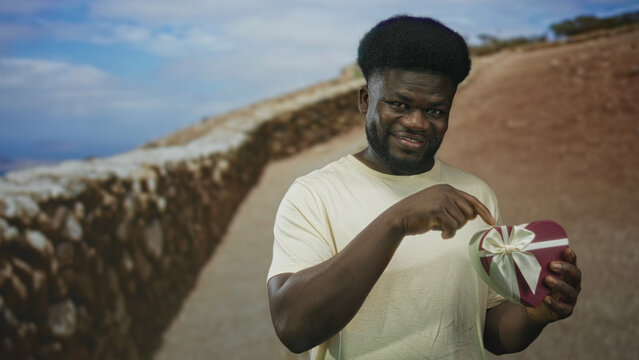 Man points finger to camera while holding heart shaped gift box with ribbon on a stone street beside a low wall; romantic surprise.