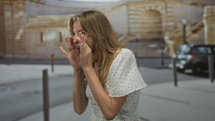 Fototapeta premium Woman cupping hands to shout on a city street beside parked car and bollards; distress urgency plea.