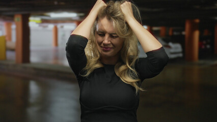 Woman smiling indoors in a dimly lit parking garage, hands in hair, wearing black outfit with a casual relaxed pose, surrounded by cars and concrete columns.