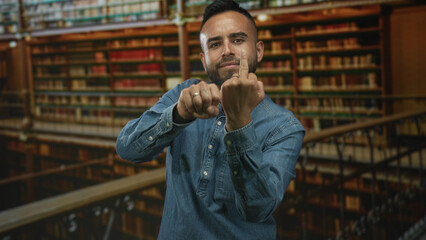 Young man in denim shirt shows middle finger gesture toward camera in a library building filled with books; defiance rebellion.