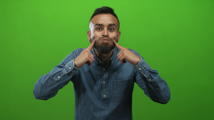 Young hispanic man with puffed cheeks and beard pointing both index fingers while wearing denim shirt in green studio; playful amusement.