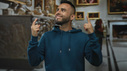 Obraz premium Man pointing both index fingers upward inside museum building with framed paintings and sculptures; playful curiosity.