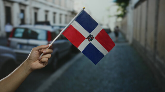 Hand holding dominican republic flag outdoor on a city street capturing vibrant cultural pride in a bustling urban town with cars and buildings in the background. - Powered by Adobe