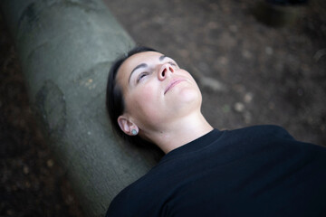 A young woman in a black T-shirt lying on a fallen tree trunk in the forest with her hand under her head, eyes open
