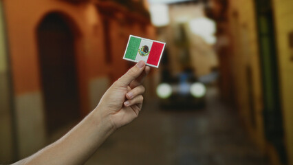 Closeup of a caucasian man's hand holding an embroidered mexican flag patch in a vibrant outdoor city street.