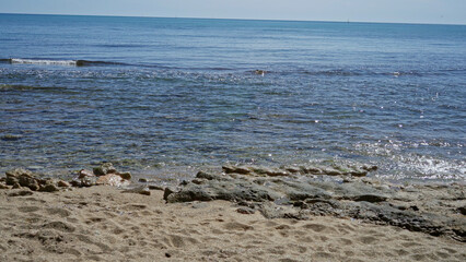 Mediterranean beach scene with clear water, sunlit sand, and gentle waves outdoors, illustrating a tranquil seaside atmosphere under a bright blue sky.