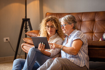 Portrait of happy mature blonde curly woman sitting on the floor close to her elderly mother sharing together social news on digital tablet. The scene captures affection, and emotional connection