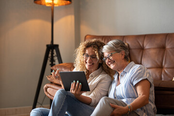 Portrait of happy mature blonde curly woman sitting on the floor close to her elderly mother sharing together social news on digital tablet. The scene captures affection, and emotional connection