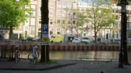 Blurred view of amsterdam canal scene with bicycles and parked cars in soft focus, characterized by green trees and urban architecture under sunny sky.