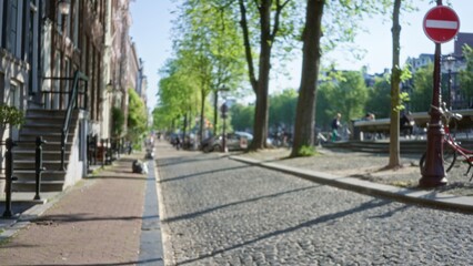 Fototapeta premium European city street with blurred perspective featuring cobblestones, trees, bicycles, and people enjoying a sunny day outdoors in an urban setting.