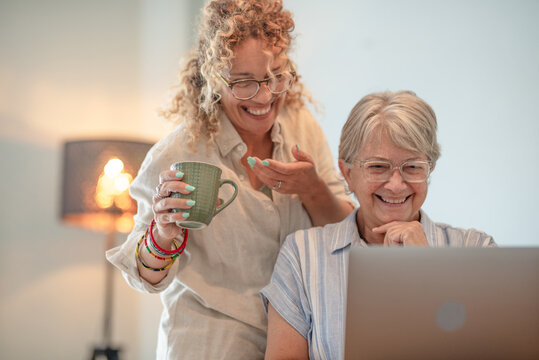 Blonde curly woman helping an older woman use a laptop at home while having a coffee break and smiling. The image represents intergenerational connection, technology learning, and family bonding