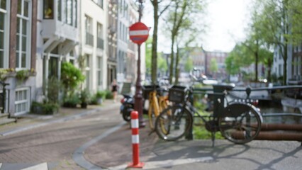 Obraz premium Defocused view of bicycles and red sign in a sunny amsterdam street with trees and canal, capturing urban tranquility and charm of a typical netherlands city scene.