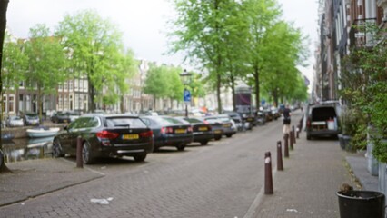 Blurred urban scene captures defocused cars parked along a picturesque canal street in amsterdam with lush green trees and a distant woman jogging, creating a serene backdrop.