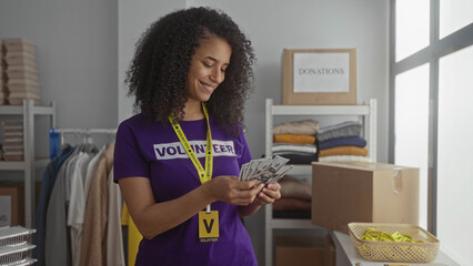 Woman counting money in charity center wearing volunteer shirt indoors with donation boxes and...