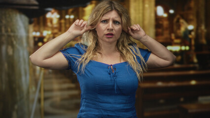Young blonde woman in blue dress pressing fingers into ears and closing eyes inside church building near wooden pews; distress isolation.