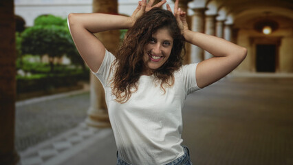Woman smiling with hands on head and fingers making bunny ears gesture in building colonnade wearing white t shirt and denim jeans; playfulness.