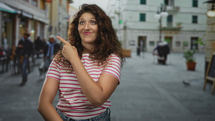 Woman with striped shirt points finger to the right, smiling and showing forearm on a busy street near cafe tables; playful curiosity.