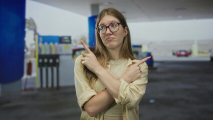 Blonde woman wearing glasses and beige jacket pointing both fingers at petrol station forecourt amid fuel pumps  indecision. © Krakenimages.com