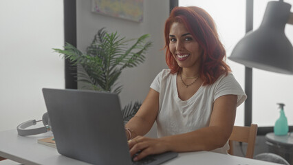 Woman typing on laptop in building home office at desk with headphones and plant visible; remote work focus.